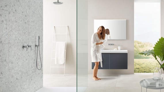 Nainen kuivaa hiuksiansa pyyhkeellä kylpyhuoneessa, jossa on avoin suihku ja suuret laatat terrazzo-tyyliin