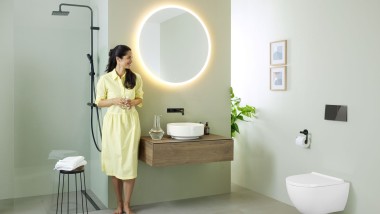Keltaiseen mekkoon pukeutunut nainen seisoo mintunvihreän kylpyhuoneen edessä, joka on varustettu Geberitin kylpyhuonekalusteilla ja kylpyhuoneposliineilla sekä mustilla hanoilla