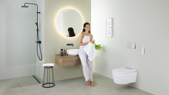 Nainen nojaamassa pesualtaaseen kylpyhuoneessa, jossa on Geberit AquaClean Sela -pesu-wc-istuin ja Geberit VariForm -pesuallas sekä huonekaluja
